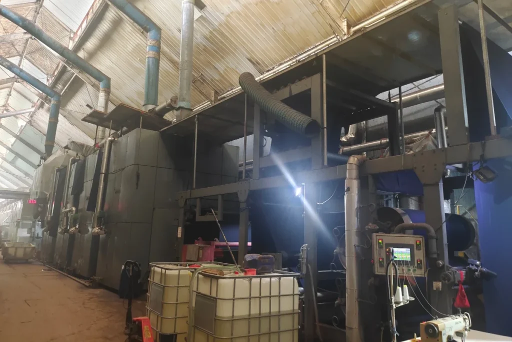Safe-Guard's ammonia curing range pre-part process: A textile web is padded with the flame retardant agent (white IBC containers in the foreground) before entering the curing chamber in our factory.